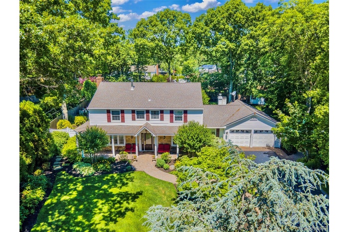 Traditional colonial two-story residence featuring light-colored siding, a covered entry porch, a multi-car garage, mature landscaping, and a paver wa