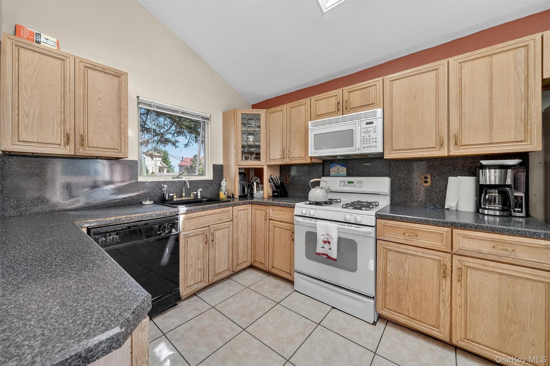 Kitchen with light brown cabinetry, white appliances, vaulted ceiling, tasteful backsplash, and light tile patterned floors