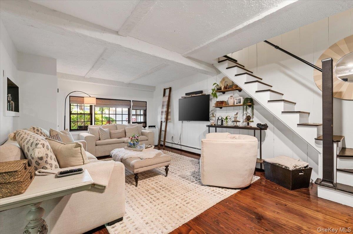 Living area featuring wood-finish flooring, exposed ceiling beams, and a modern staircase with dark treads and a black handrail