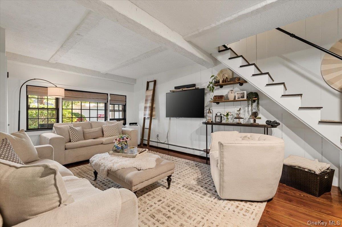 Open-concept living area featuring exposed ceiling beams, wood-finish flooring, and a staircase with painted risers