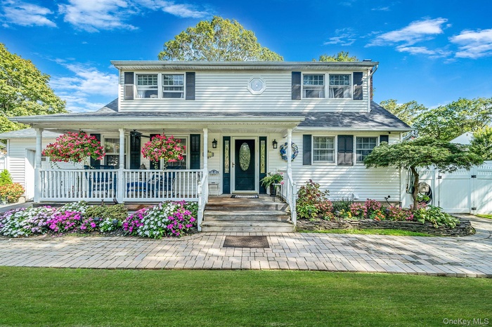 White siding exterior featuring a covered front porch with white railings, a paver walkway, mature landscaping, and a dark front door with decorative 