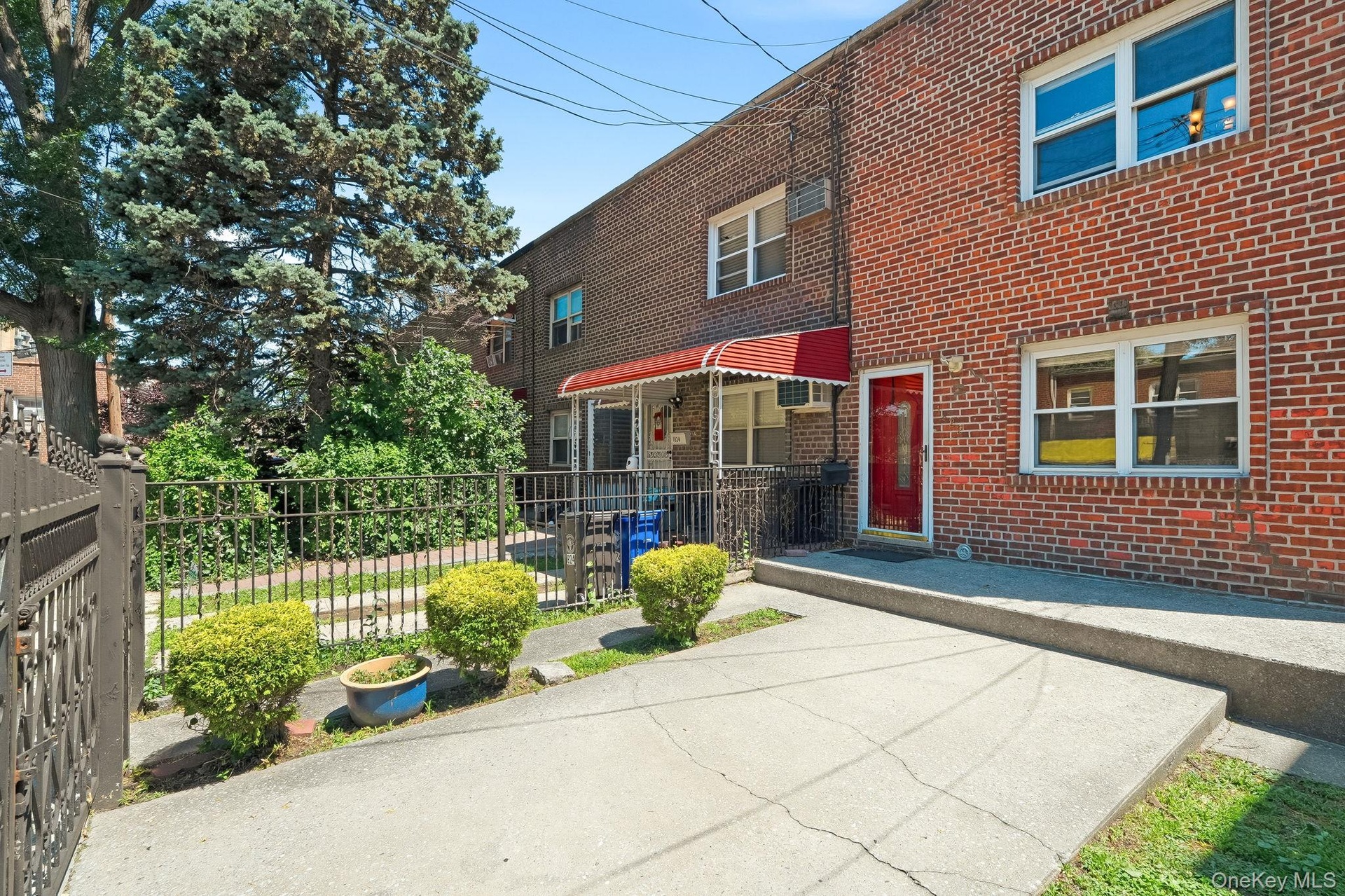 Brick facade exterior featuring a red entry door, a red striped awning, a concrete driveway, and a black wrought iron fence.