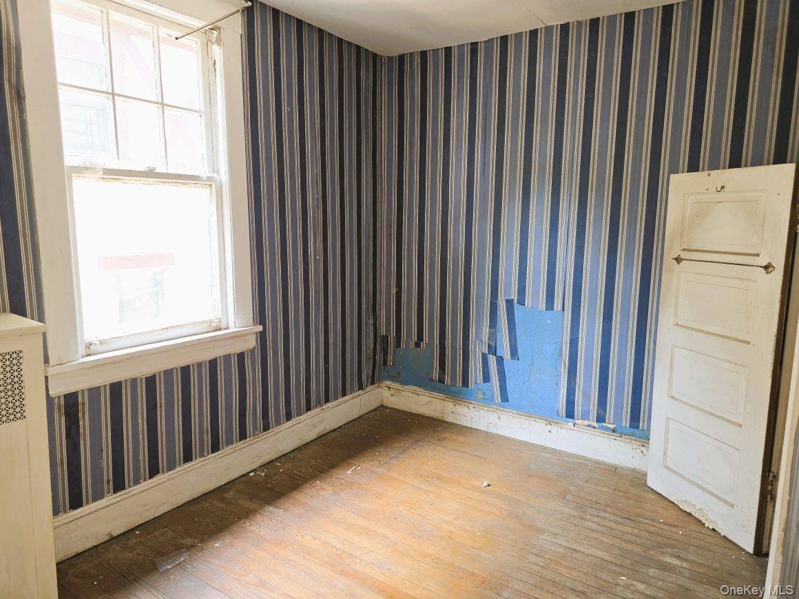 Room featuring wood-finish flooring, striped wallpaper, a double-hung window with white trim, a white baseboard, and a white interior door