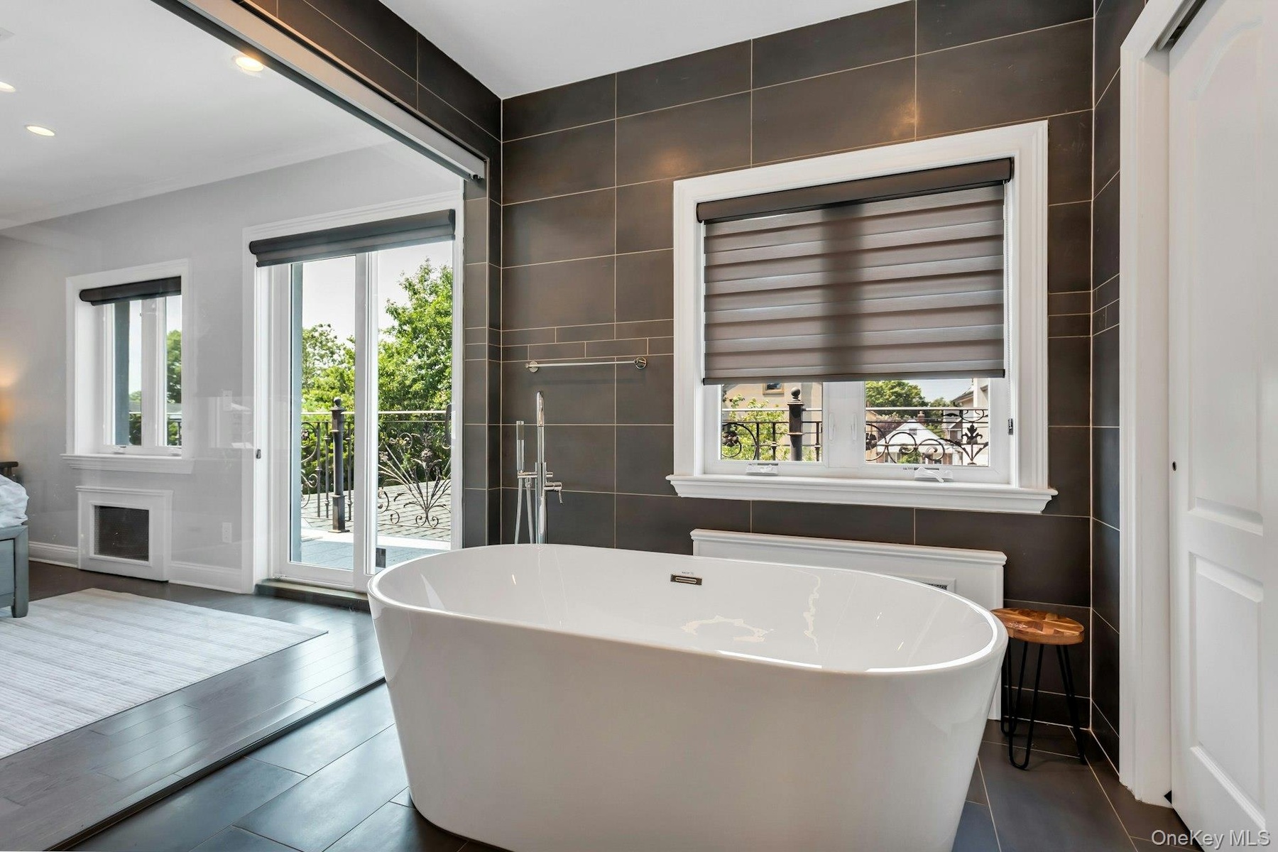Architectural Digest! Freestanding soaking tub with floor-mounted faucet, dark large-format tile accent wall, dark wood-finish flooring, and a glass-p
