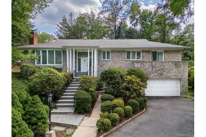 Stone facade residence featuring a prominent front entrance with white columns, a two-car garage, and extensive mature landscaping.