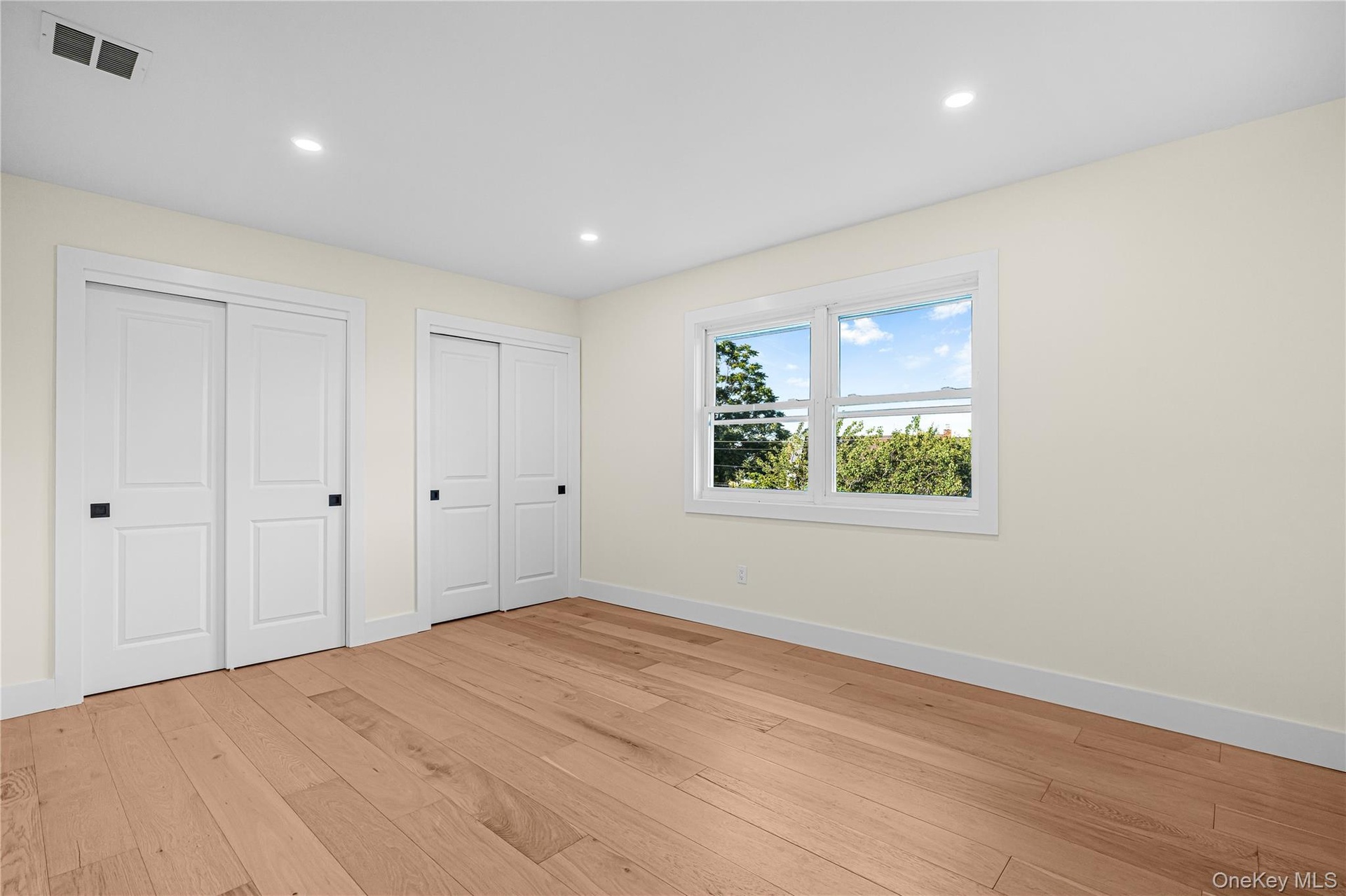 Spacious room featuring wood-finish flooring, two sets of bi-fold closet doors with black hardware, a large window, recessed lighting, and white baseb