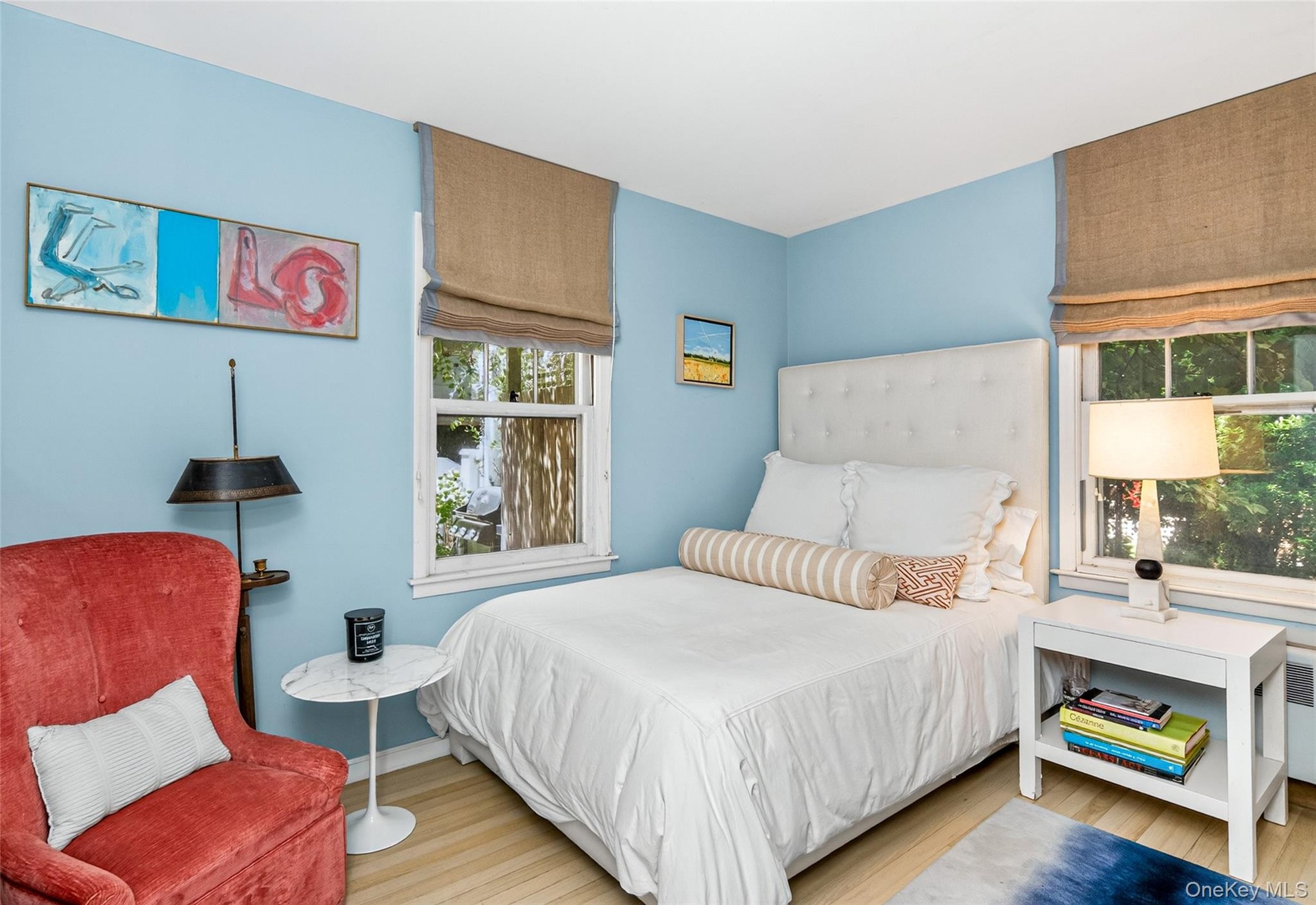 First Floor Bedroom featuring light wood-finish flooring, two large windows with natural fiber window treatments, a tufted white headboard, and light 
