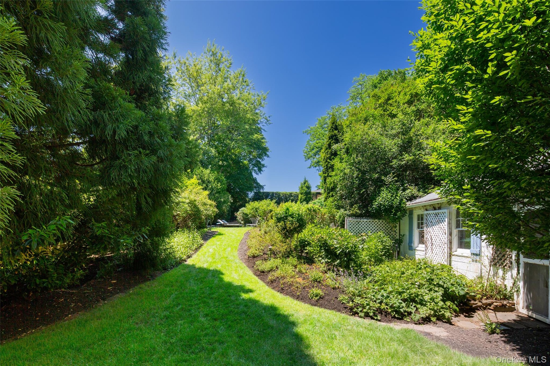 Landscaped grounds featuring a winding grassy path, mature trees, dense shrubbery, a white lattice fence, and a white shed with blue shutters