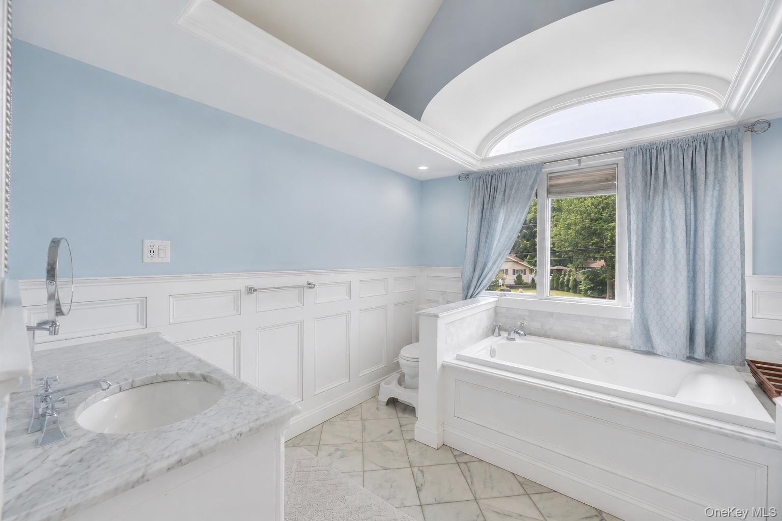 Bathroom featuring a spacious built-in soaking tub, an arched transom window, a single vanity with a stone countertop and undermount sink, wainscoting