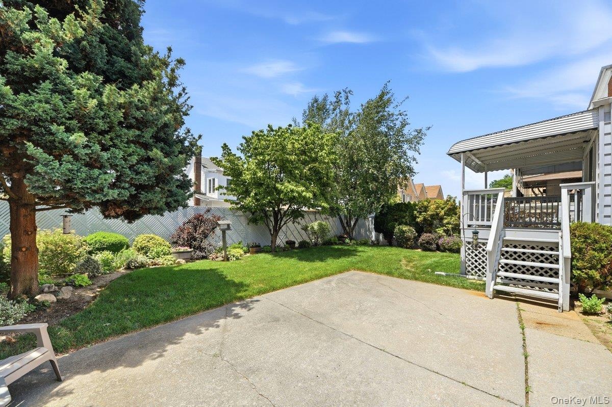 Manicured backyard with concrete patio, lush lawn, mature trees, and a covered porch with white railings and lattice detailing