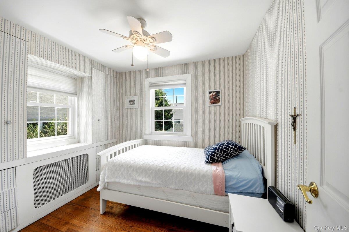 Hardwood flooring grounds this first floor bedroom, featuring two windows with white trim, a built-in radiator cover