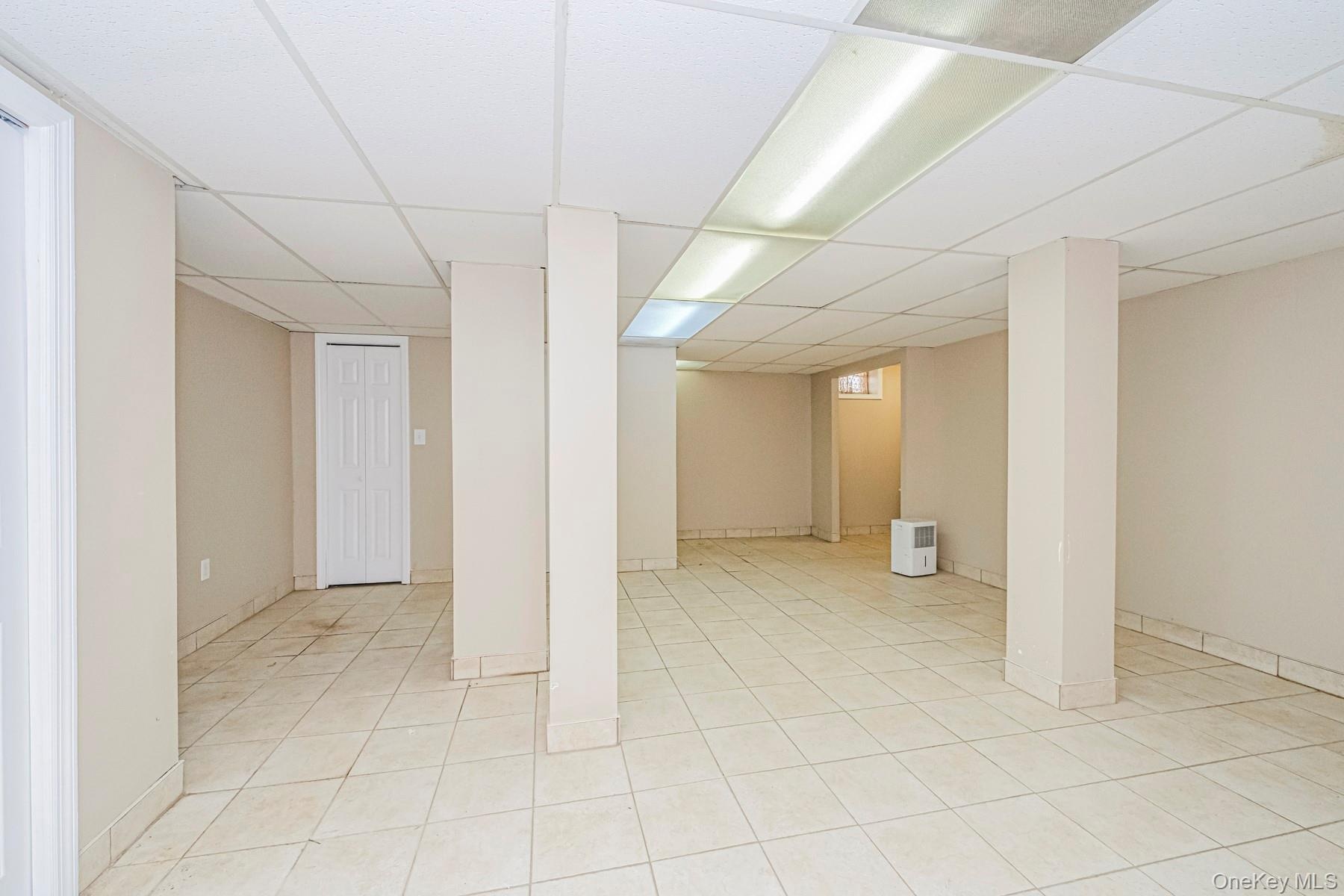 Spacious room featuring light-toned tile flooring, neutral wall paint, and a drop ceiling with integrated lighting
