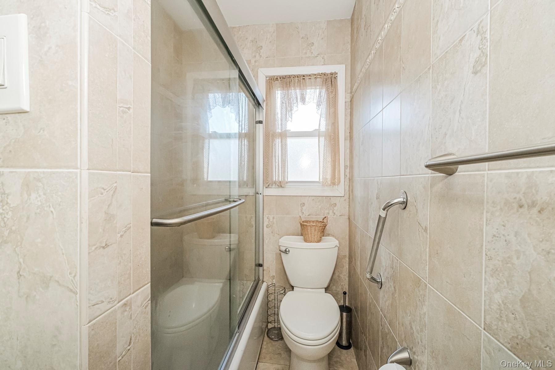 Bathroom featuring floor-to-ceiling ceramic tile, a glass-enclosed shower-tub combination, and a window with frosted privacy glass