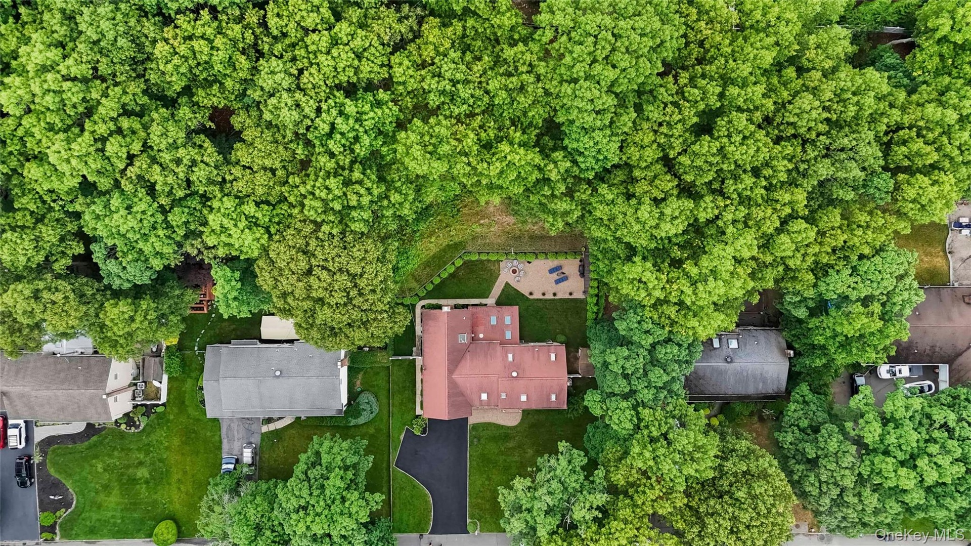 Property surrounded by mature trees, featuring a red-toned roof, multiple skylights, and an expansive paved driveway