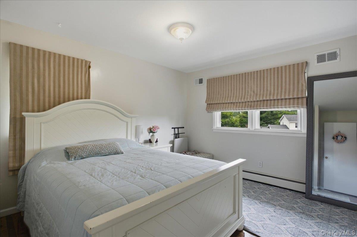 Spacious bedroom featuring light-toned walls, wood-finish flooring, a contemporary ceiling light fixture, and a window with a striped Roman shade