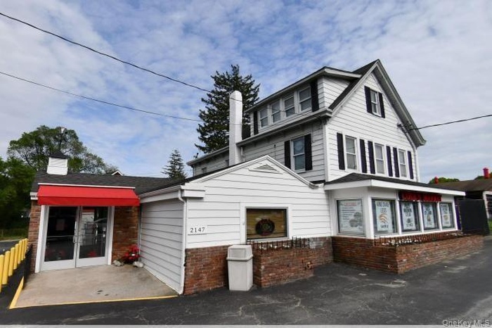 Two-story building featuring white siding, multiple windows with dark shutters, and a prominent red fabric awning over an entrance