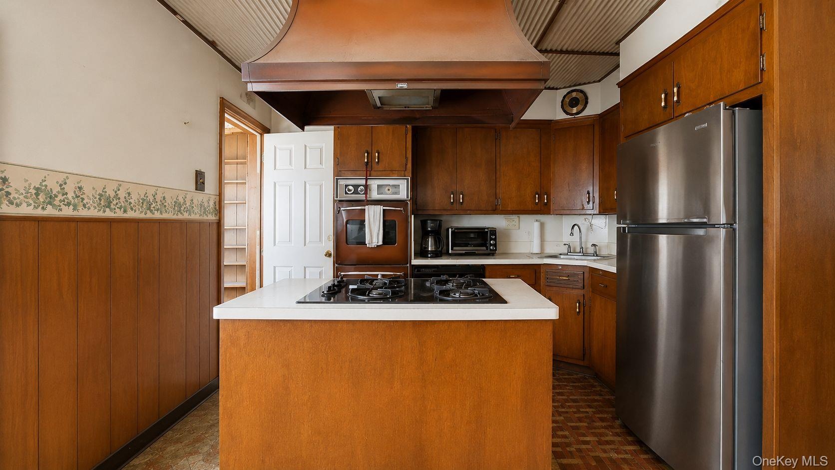 Kitchen featuring wood cabinetry, stainless steel refrigerator, white countertops, built-in oven, and a center island with cooktop
