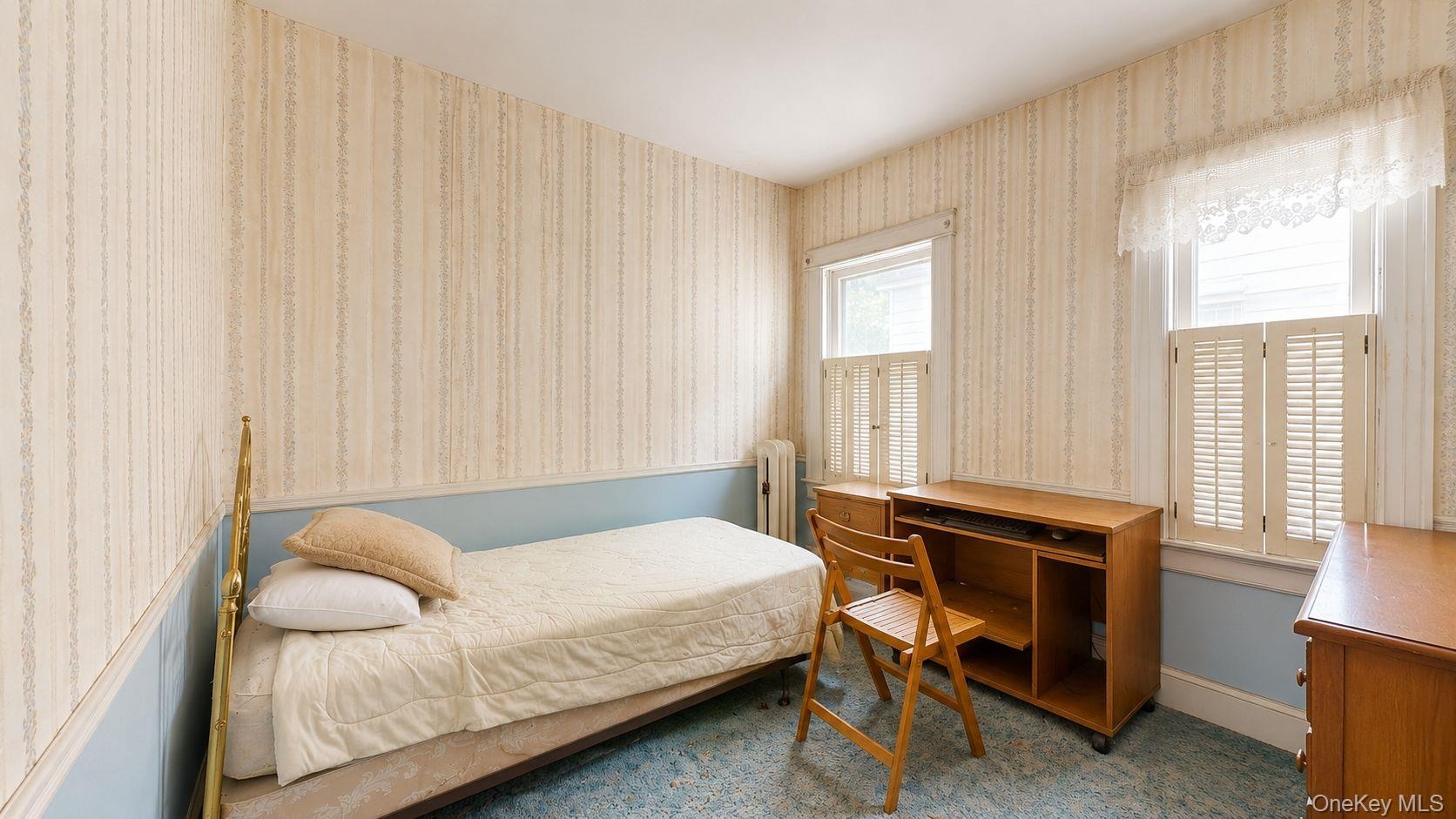Bedroom featuring two windows with white louvered shutters, a built-in radiator, and light-toned patterned wallpaper with a blue wainscoting detail
