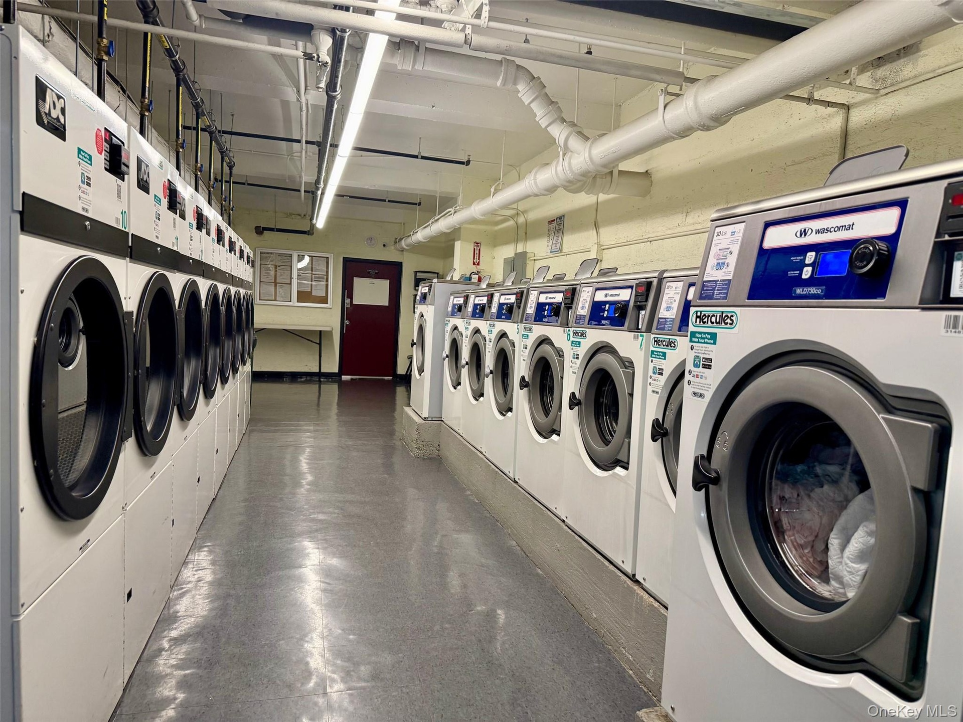 Community laundry room featuring multiple front-loading washing machines, durable flooring, and overhead fluorescent lighting