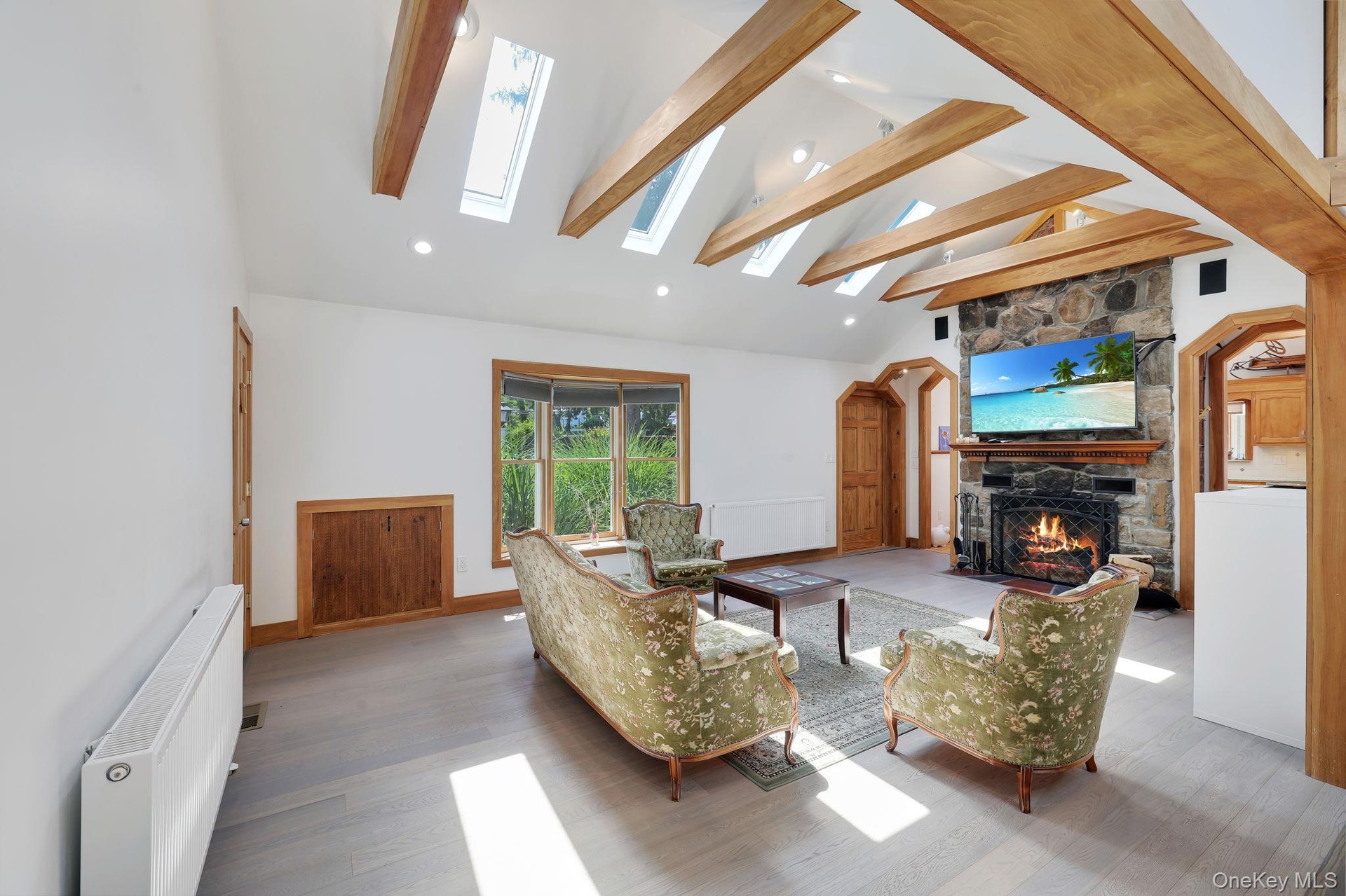 Spacious living room featuring a stone fireplace, a vaulted ceiling with exposed wood beams, multiple skylights, and light-toned wood-finish flooring.