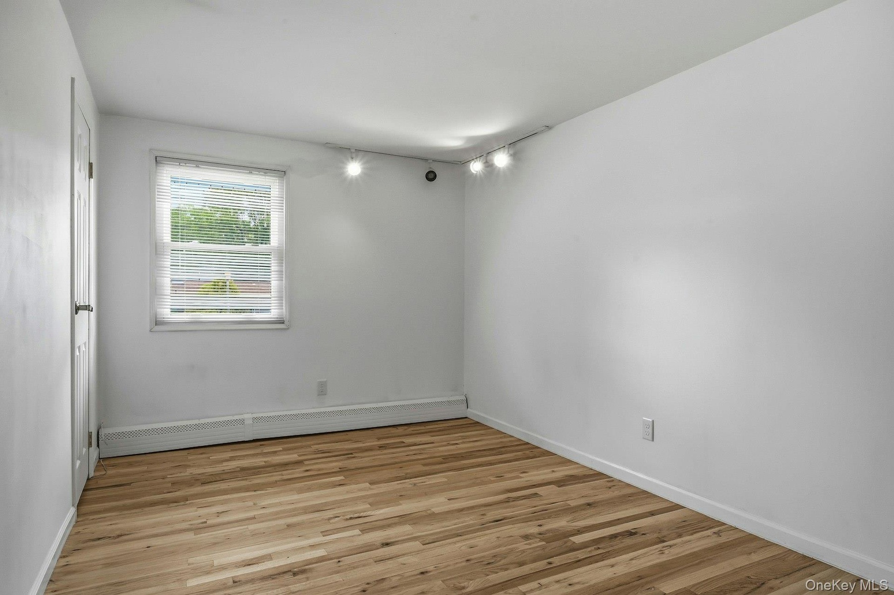 Room featuring wood-finish flooring, white painted walls, a single window with blinds, a baseboard heater, and ceiling-mounted track lighting