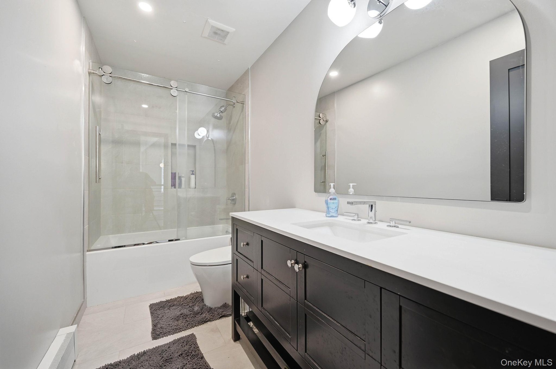 Modern bathroom featuring a dark wood-finish vanity with a white countertop and undermount sink, a large arched mirror, and a tub/shower combination w