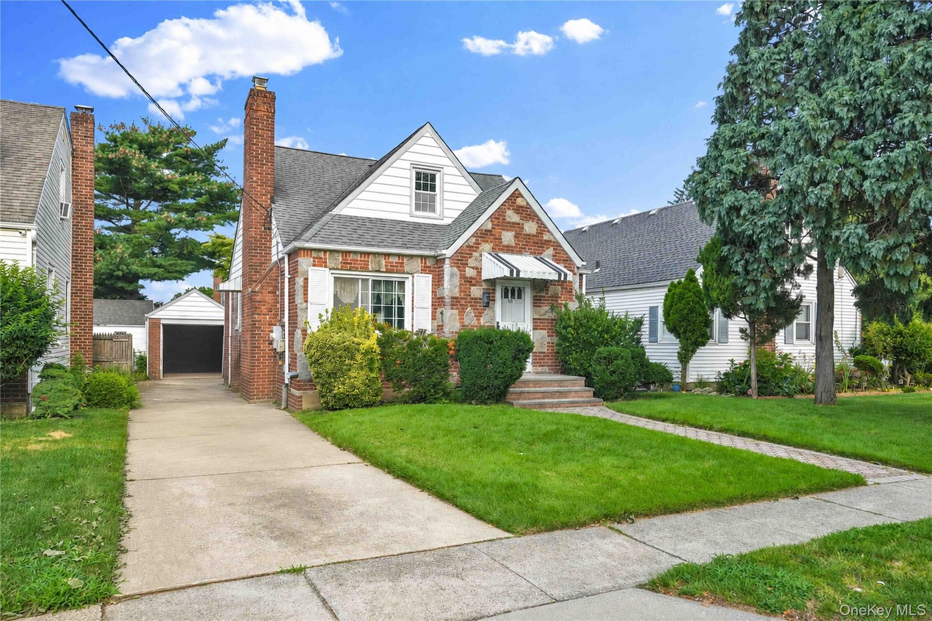 Charming brick Colonial with crisp white trim and peaked roofline, featuring a manicured front lawn, established landscaping with mature shrubs, concr