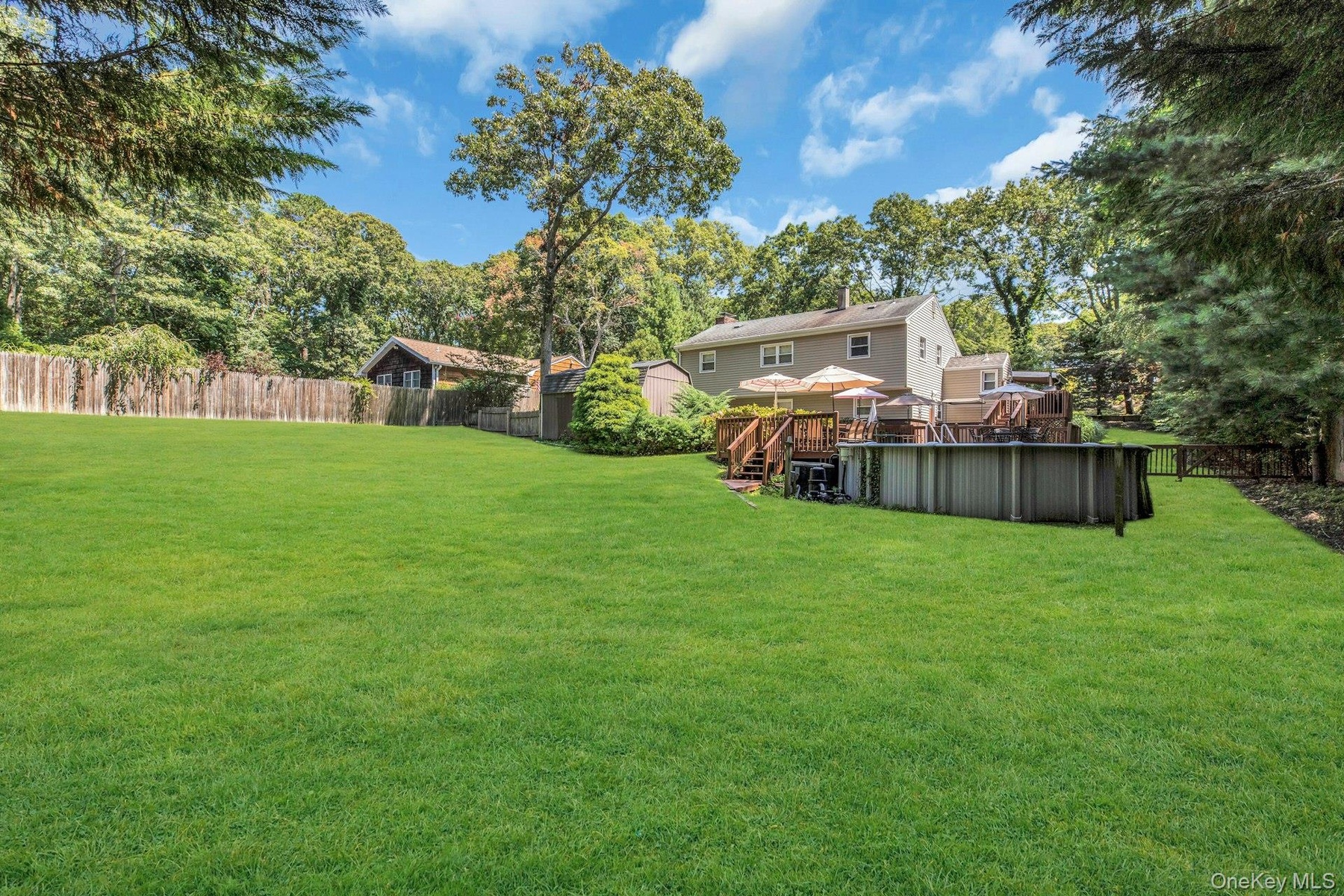 Expansive manicured lawn with mature tree canopy, wooden deck with integrated seating, detached storage structure, and fenced perimeter