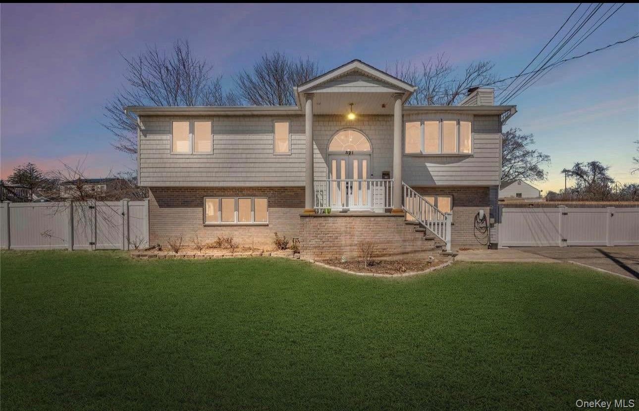 Elevated colonial-style home featuring a manicured lawn, vinyl siding exterior, brick foundation detailing, white vinyl fencing, and illuminated front