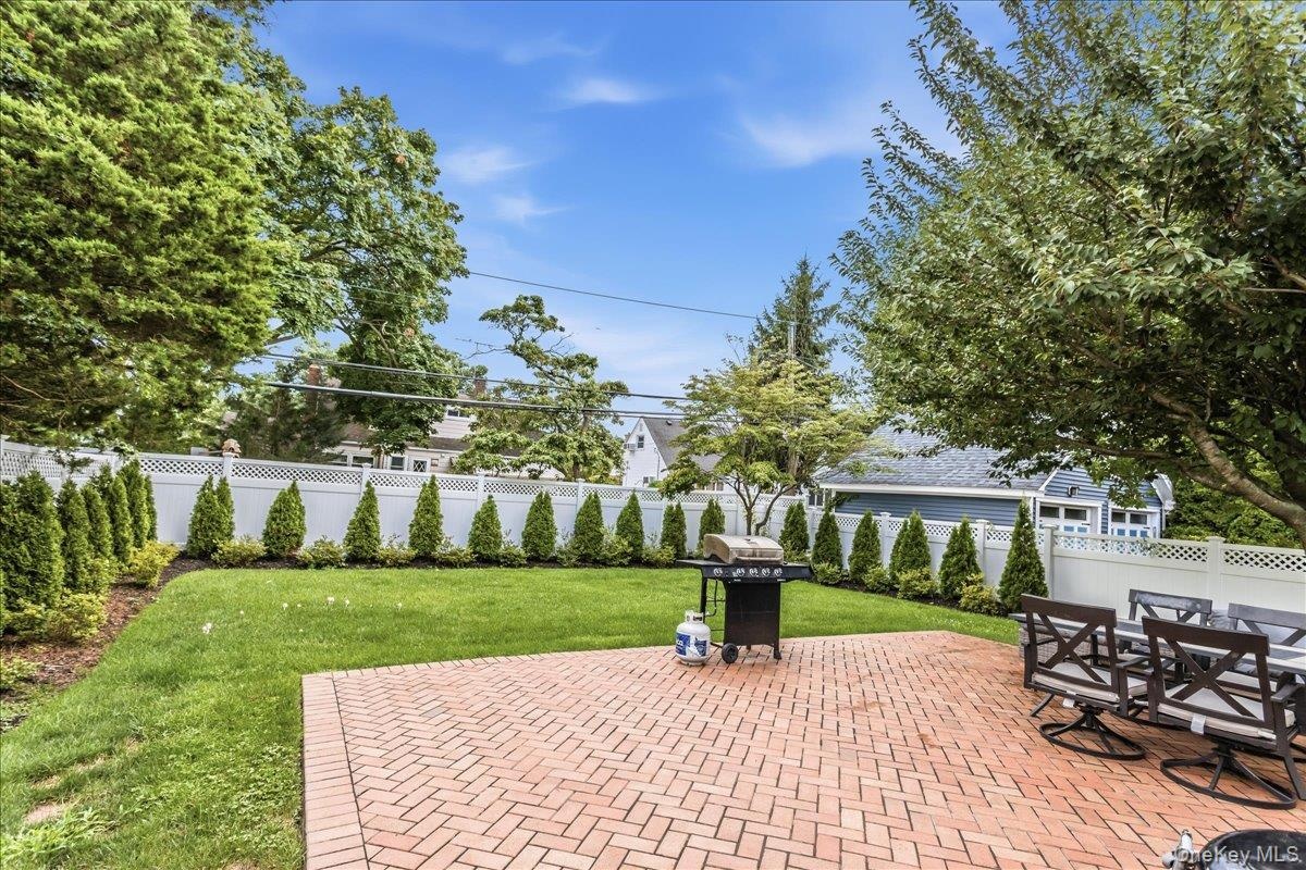 Expansive backyard featuring a brick paver patio, a manicured lawn, a white lattice fence, mature trees, and evergreen landscaping