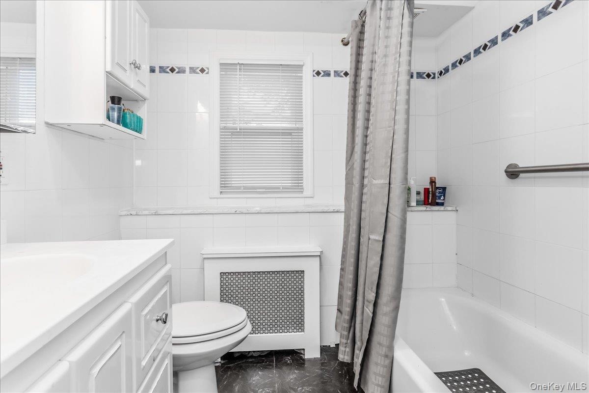 Bathroom featuring white wall tiling with decorative accent borders, a built-in bathtub with shower, a window with blinds, and a vanity with integrate