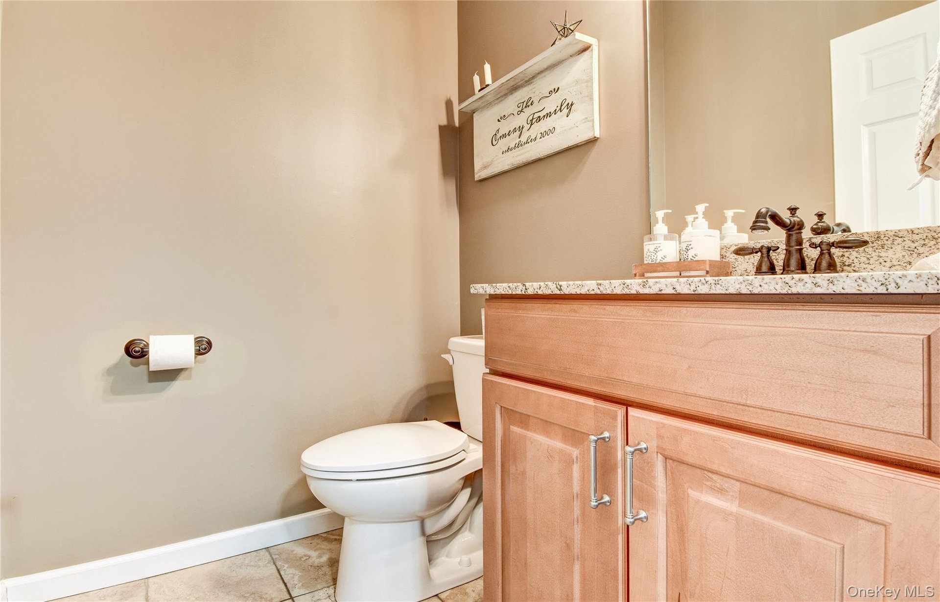 Bathroom featuring a light wood-finish vanity with paneling and brushed nickel hardware, a granite countertop, and a built-in sink with a dark-toned f