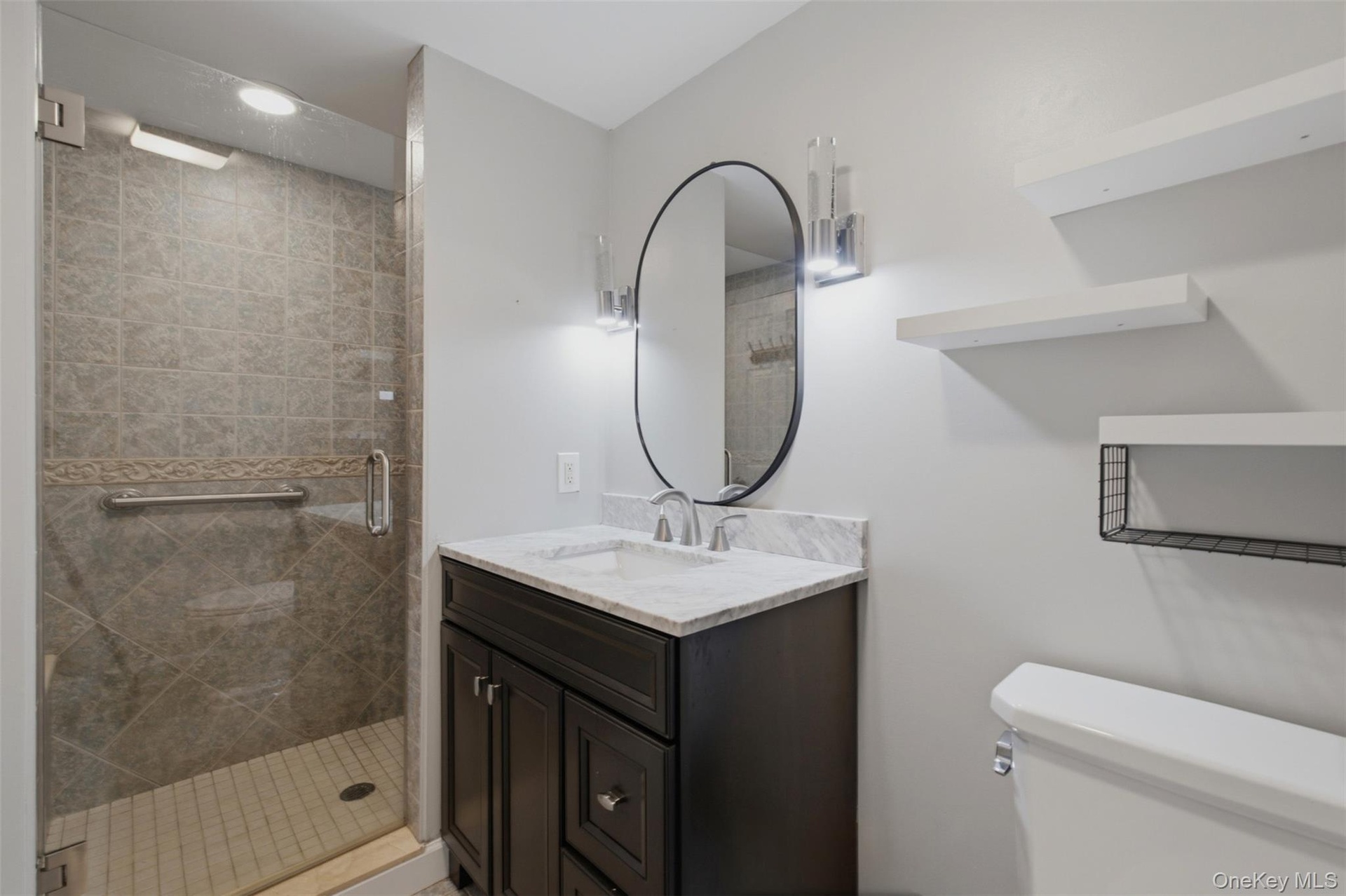 Bathroom featuring a glass-enclosed shower with tile surround, dark wood-finish vanity with a light-toned countertop, oval mirror, and wall-mounted sh