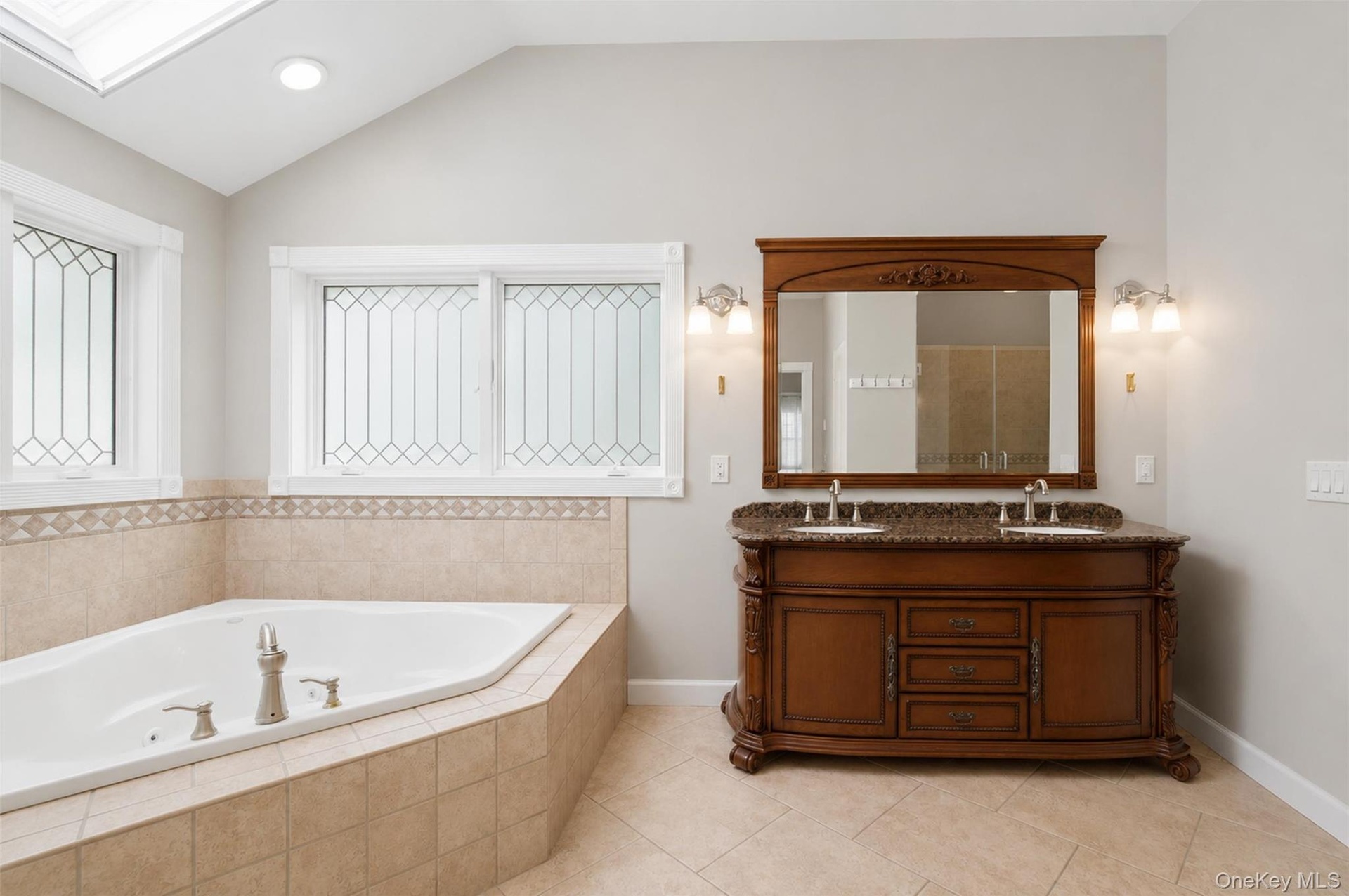 Soaking jet tub with tiled surround, dual-sink vanity with stone countertop, decorative windows, and vaulted ceiling with recessed lighting and skylig