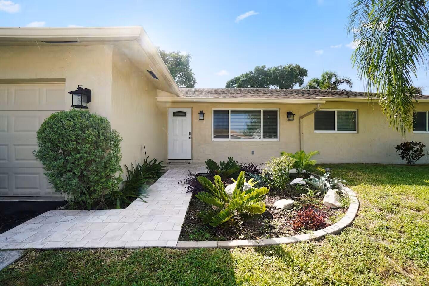 Stucco exterior featuring a tiled walkway, landscaped garden bed, white entry door with frosted glass insert, and a garage with overhead door