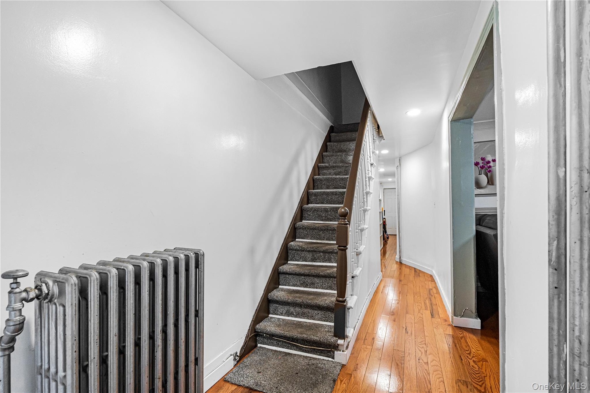 Hallway featuring hardwood flooring, a carpeted staircase with a white spindle banister, recessed lighting, and a cast iron radiator