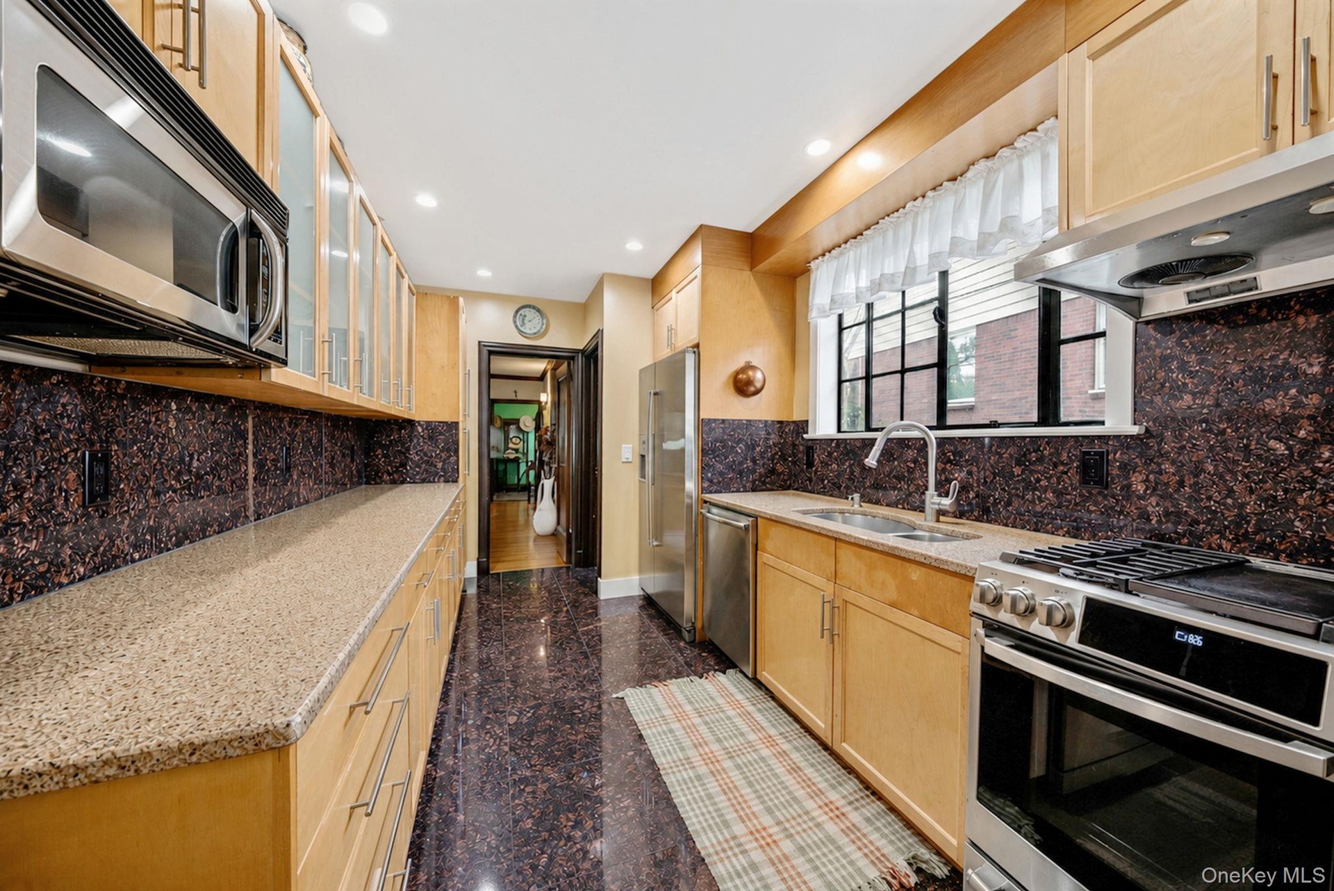Galley kitchen featuring light wood cabinetry, stone countertops, stainless steel appliances, and a dark stone backsplash