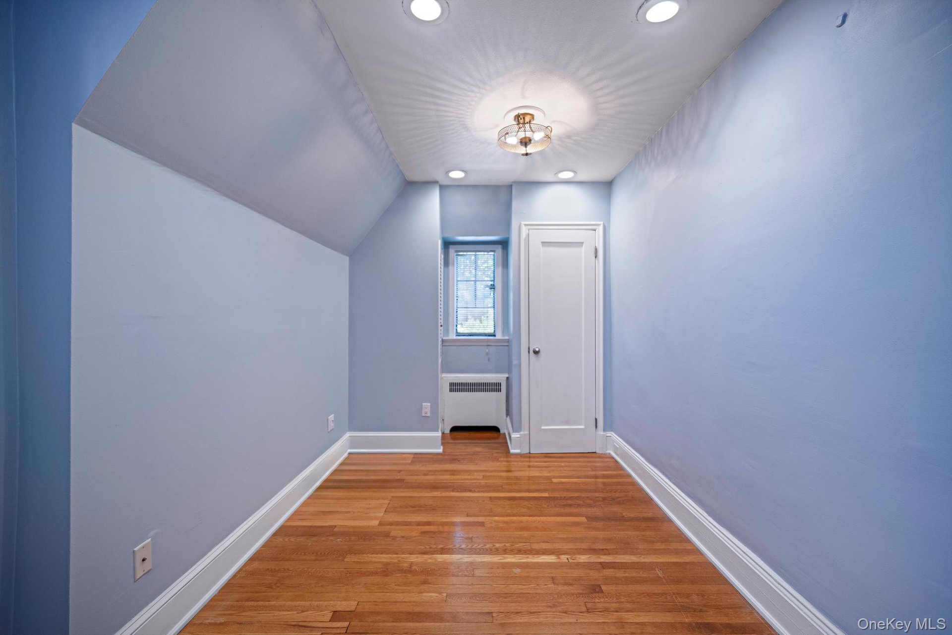 Hardwood flooring and recessed lighting illuminate this interior space, featuring a window with a radiator below