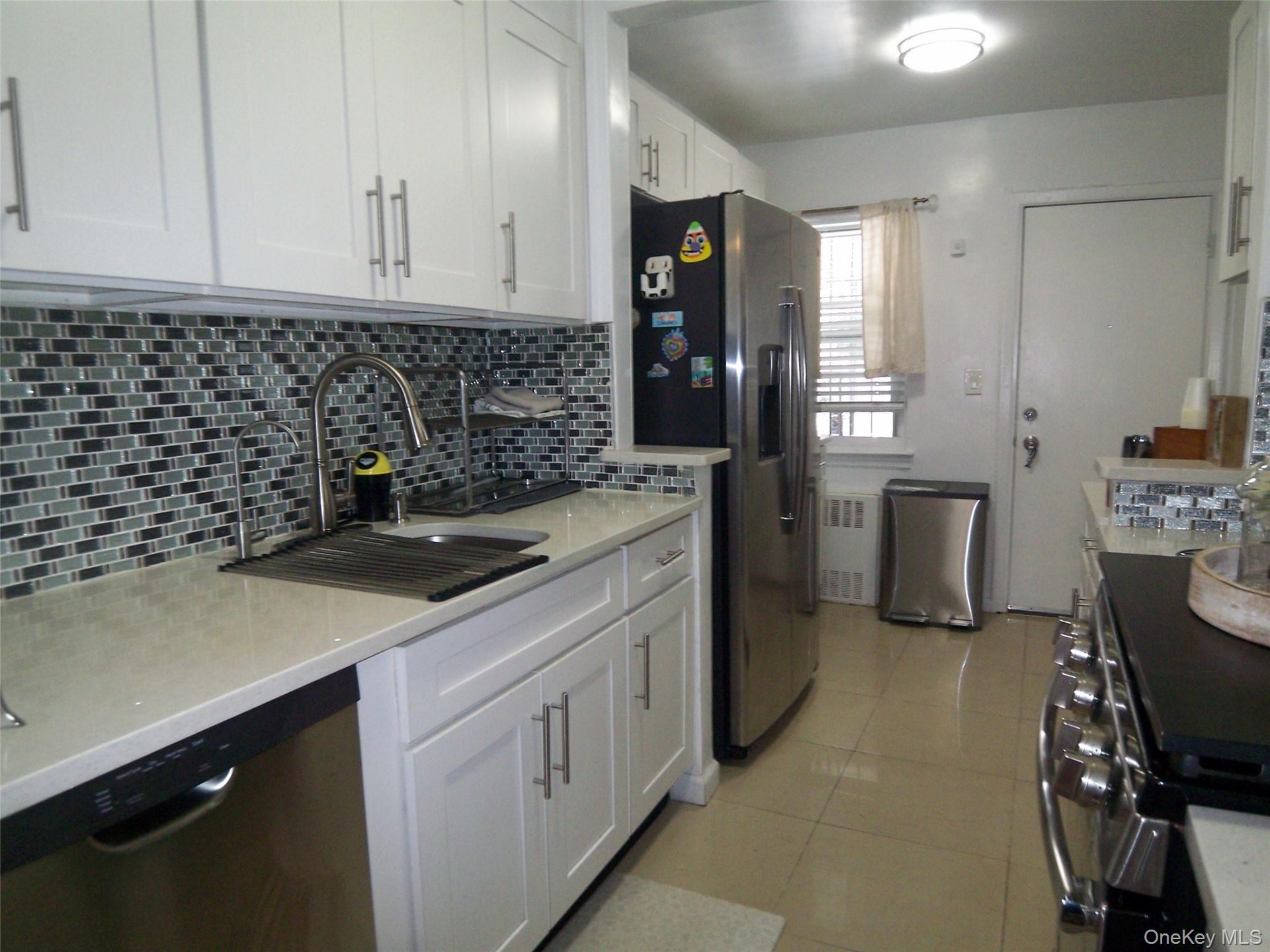 Modern kitchen featuring white shaker cabinetry, light-toned countertops, a mosaic tile backsplash, stainless steel appliances, and tile flooring