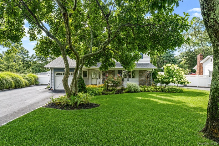 Manicured front lawn with mature shade trees, paved driveway, stone facade accents, white picket fencing, and a garage with a carriage-style door