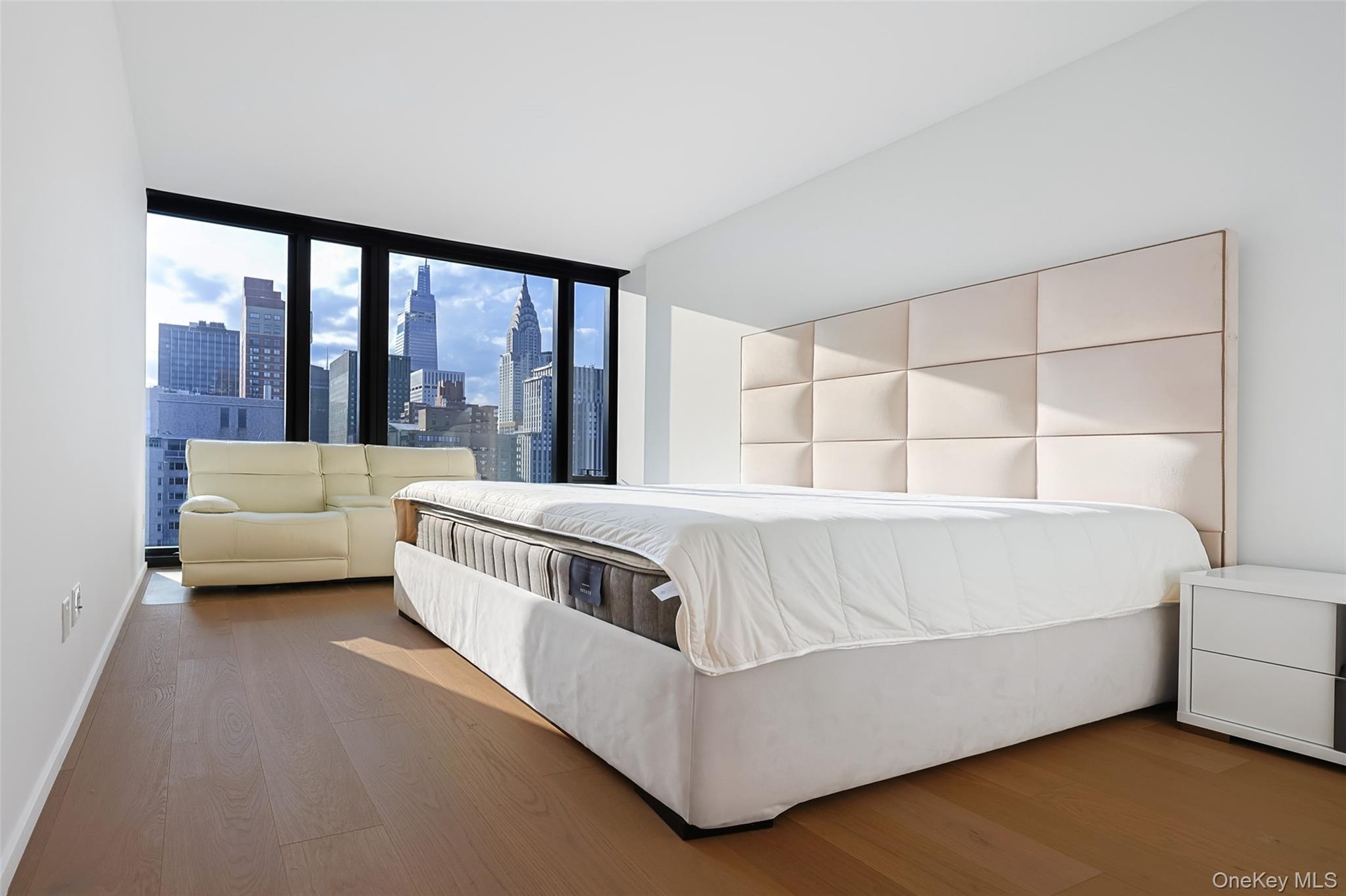 Primary Bedroom with Expansive windows showcasing cityscape views, light wood-finish flooring, white walls, and a paneled upholstered headboard