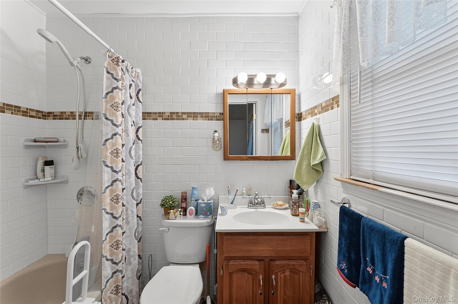 Bathroom featuring white subway tile walls with a decorative mosaic accent band, a built-in bathtub with shower, a single vanity with wood-finish cabi