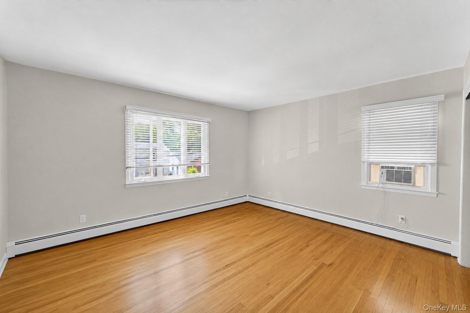 Spacious interior room featuring wood-finish flooring, light-toned walls, two windows with blinds, and baseboard heating