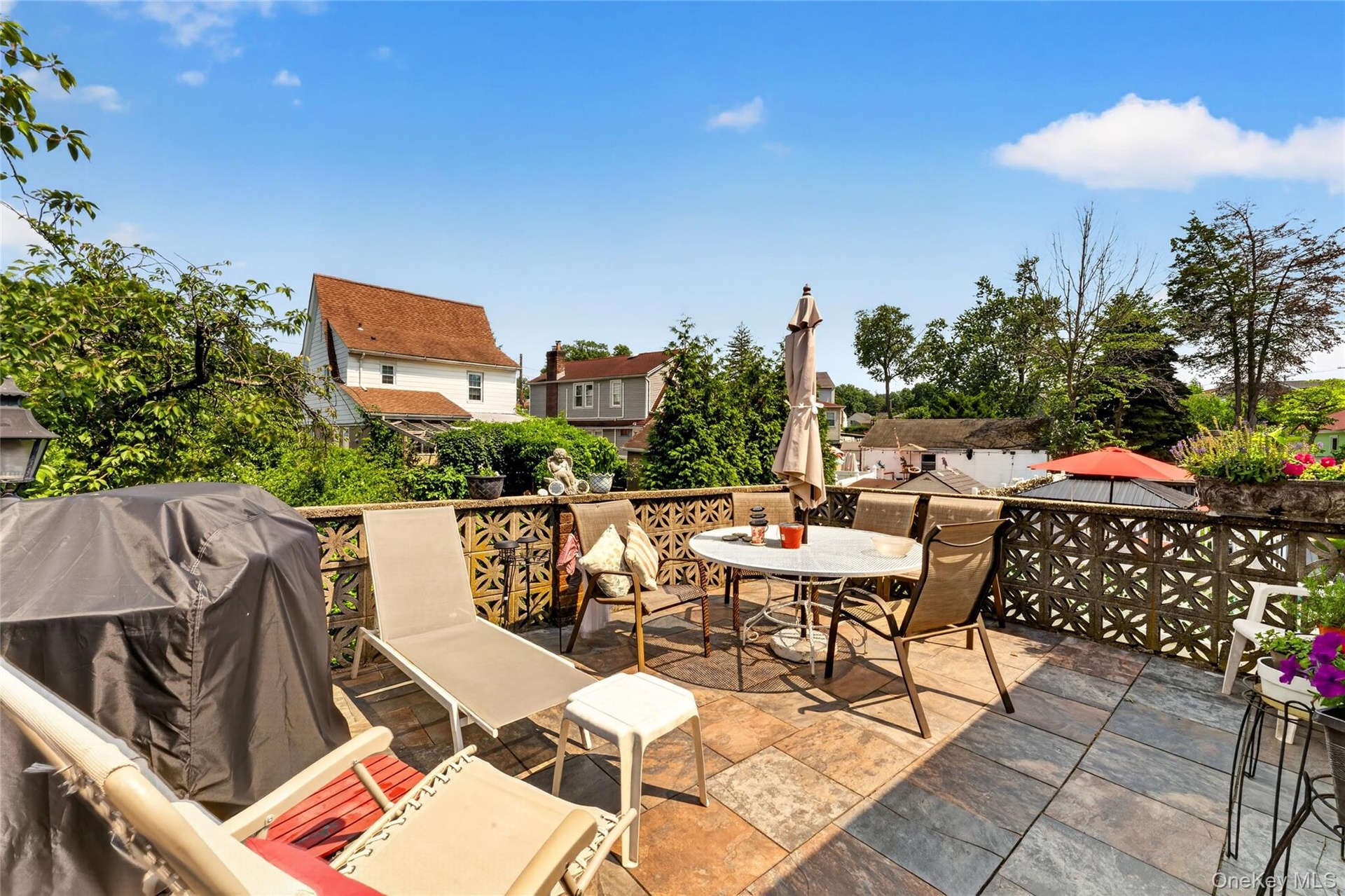 Spacious outdoor patio featuring multi-toned stone tile flooring, an openwork geometric pattern railing, and mature green foliage