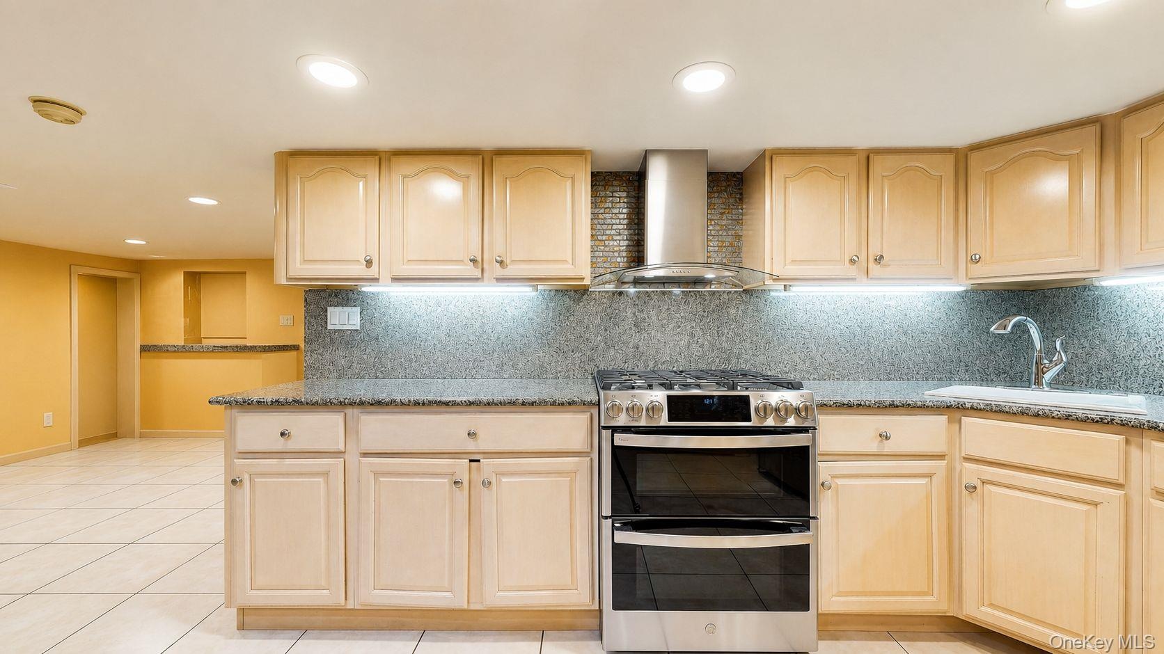 Kitchen featuring light wood-finish cabinetry, stone countertops, a stainless steel range with double ovens, a stainless steel range hood, and an unde