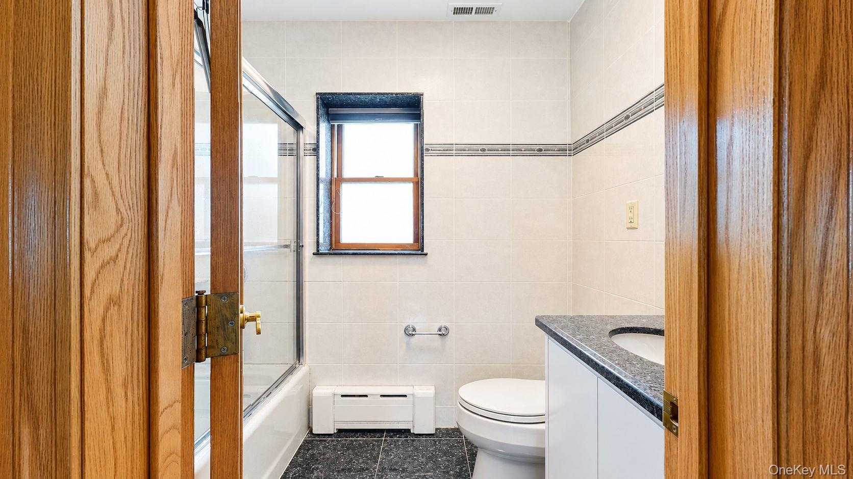 Bathroom featuring a white vanity with a dark speckled countertop, white tiled walls with a decorative border, black tile flooring, and a bathtub with