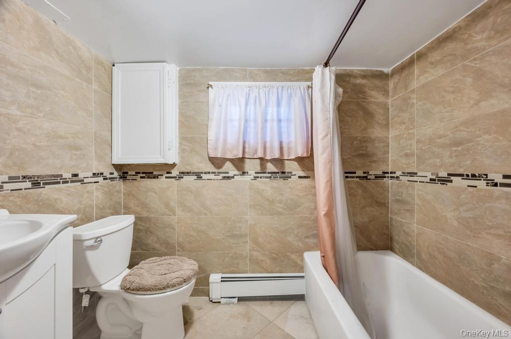 Bathroom featuring floor-to-ceiling tile, decorative mosaic accent band, white wall-mounted cabinet, window, and a built-in bathtub with shower