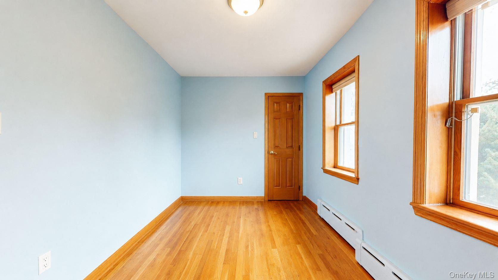 Room featuring wood-finish flooring, two windows with natural wood trim, a natural wood paneled door, baseboard heating, and a flush-mount ceiling lig