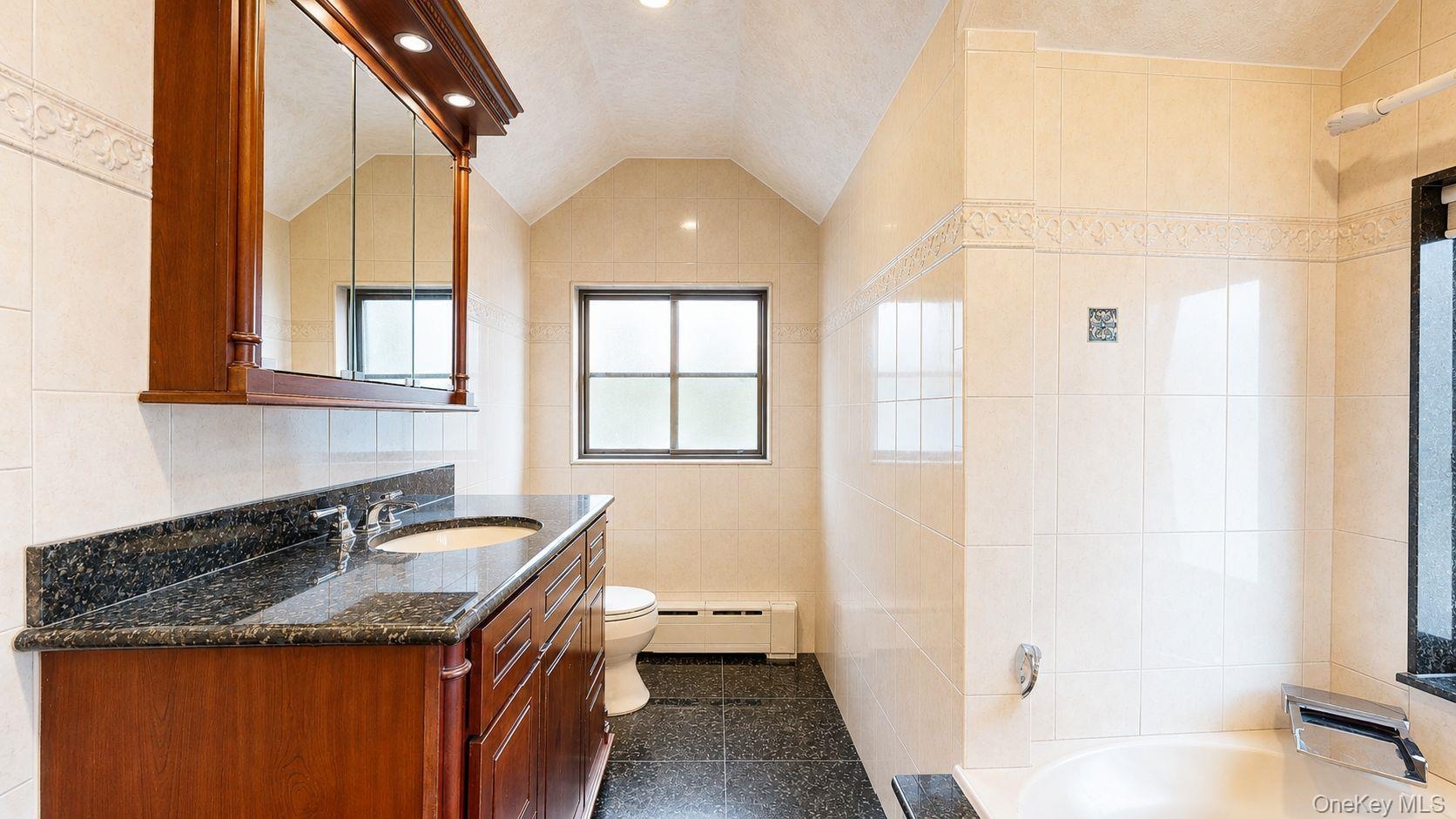 Bathroom featuring a dual-sink vanity with a dark countertop and wood-finish cabinetry, tiled walls, a bathtub with a dark surround, and a frosted win