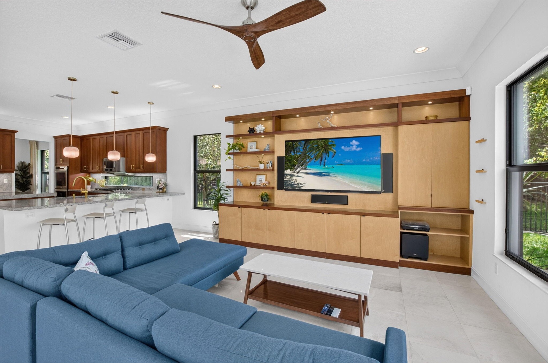Open concept living area featuring a custom media built-in with integrated lighting, wood-finish ceiling fan, and large windows